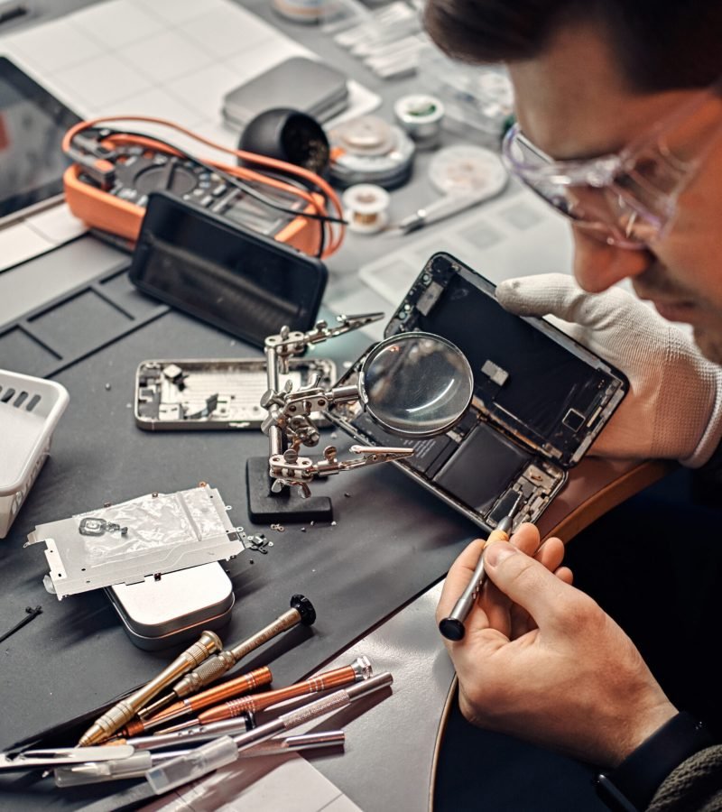 Serviceman uses magnifier and screwdriver to repair damaged smartphone in the electronic workshop.